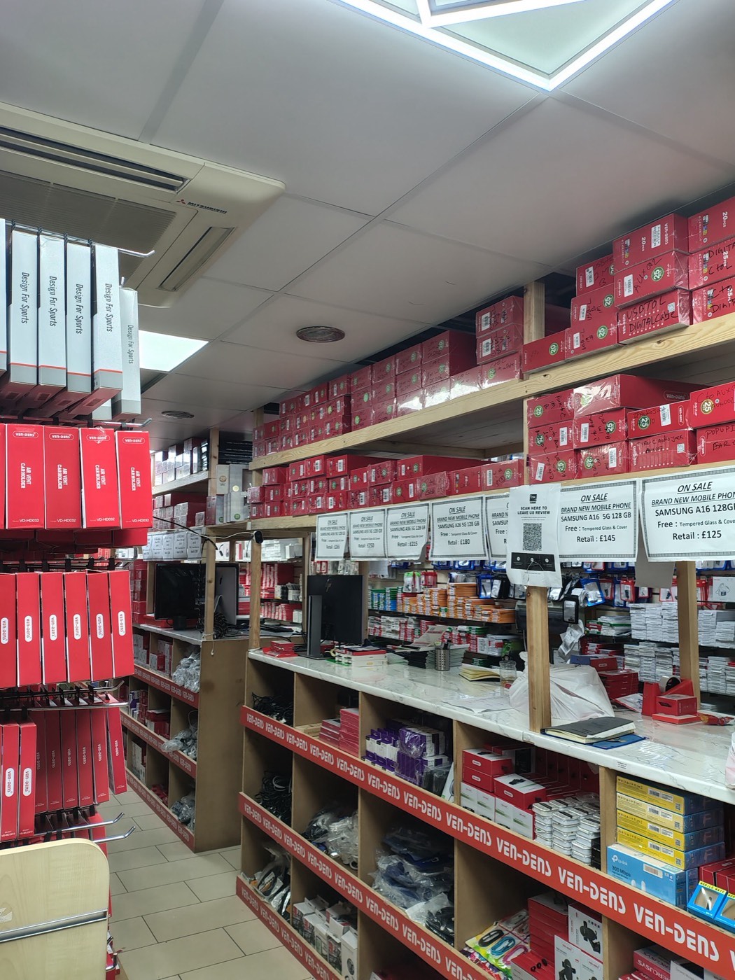 Inside Techmart looking down the aisle towards the counter, shelves stacked with boxed accessories and new mobile phones on sale.